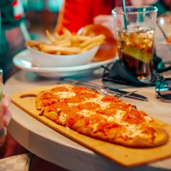 Flatbread pizza on wooden board, fries in white bowl, and soda on a table at Sports & Social at Live! at the Pointe Orlando.